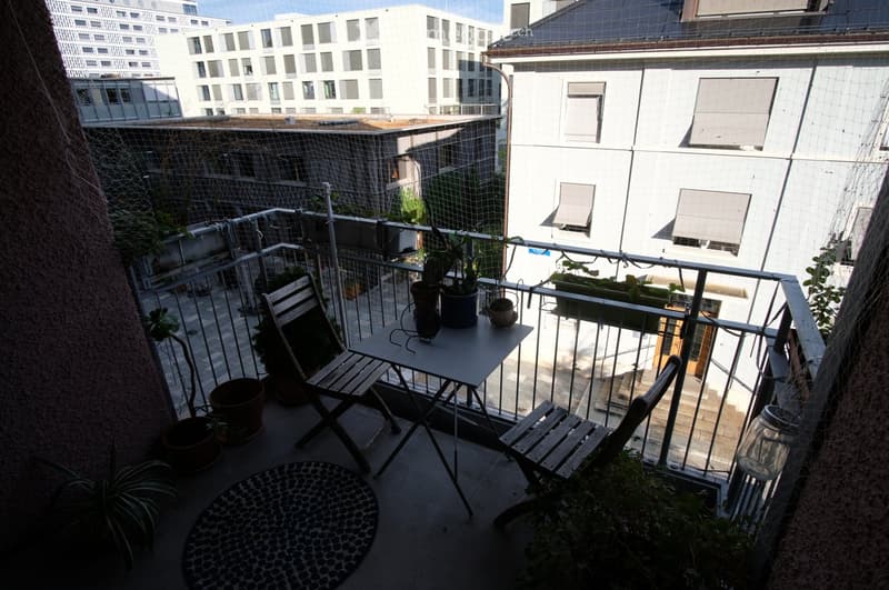 Balcony with a small table, two chairs, and several potted plants, overlooking a cityscape with modern buildings.