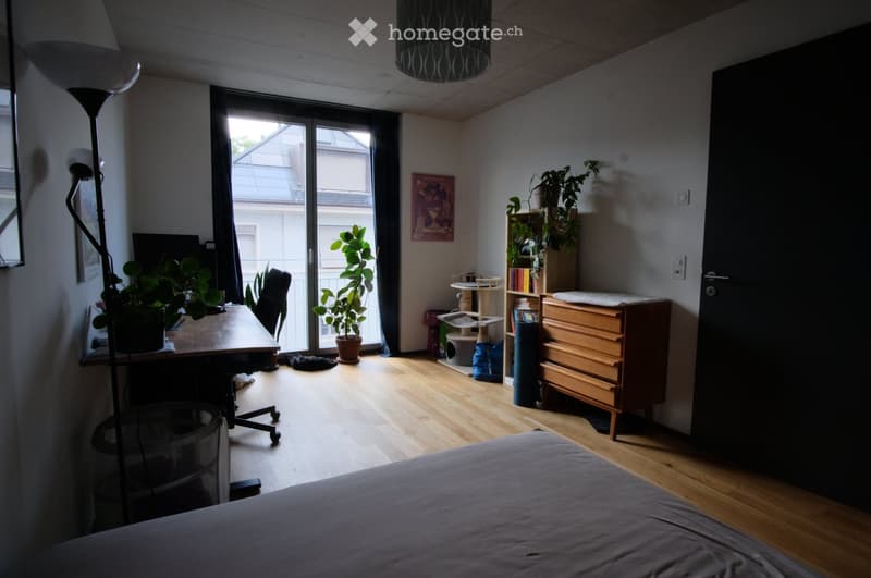 Bright bedroom with wooden flooring, large window with sliding glass door leading to balcony, bed, desk with chair, bookshelf, cat tree, potted plants, and floor lamp.