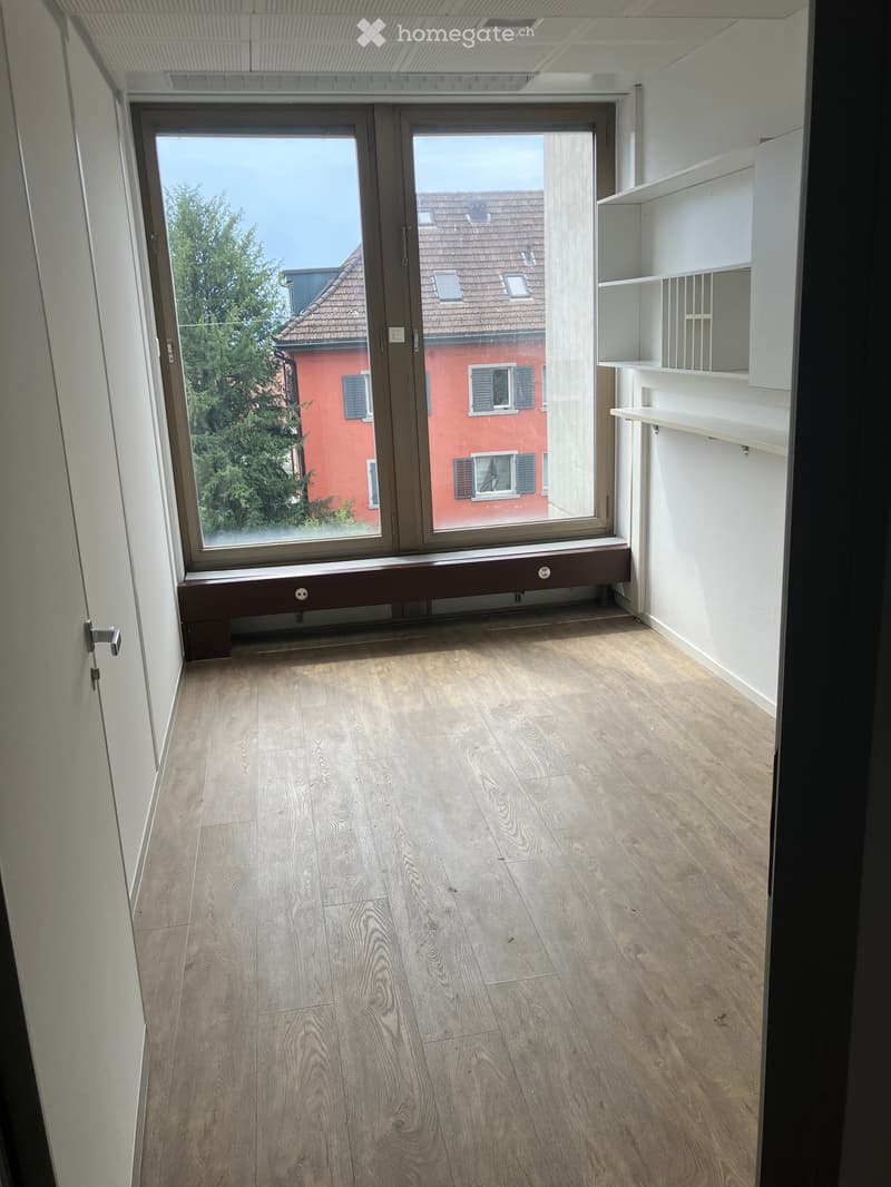 Empty room with light wooden flooring, white walls, and a large window with a view of a red building and tree. Shelves are mounted on the right wall.