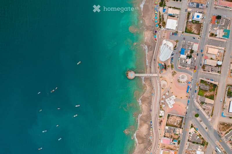 Aerial view of a coastal area with a sandy beach, turquoise water, small boats, and scattered buildings and structures along the shoreline.