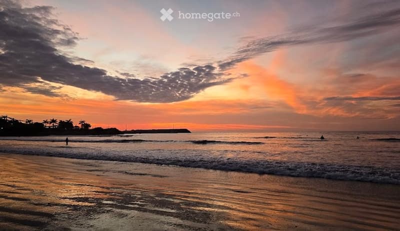 A serene beach scene at sunset with calm ocean waves, a sandy shore, and a few people enjoying the water.