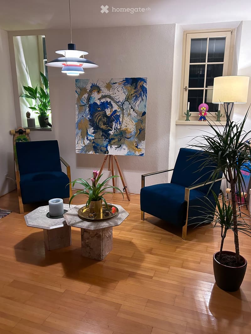 Modern living room with wooden flooring, two blue armchairs, a marble coffee table, a floor lamp, and a painting on the wall.