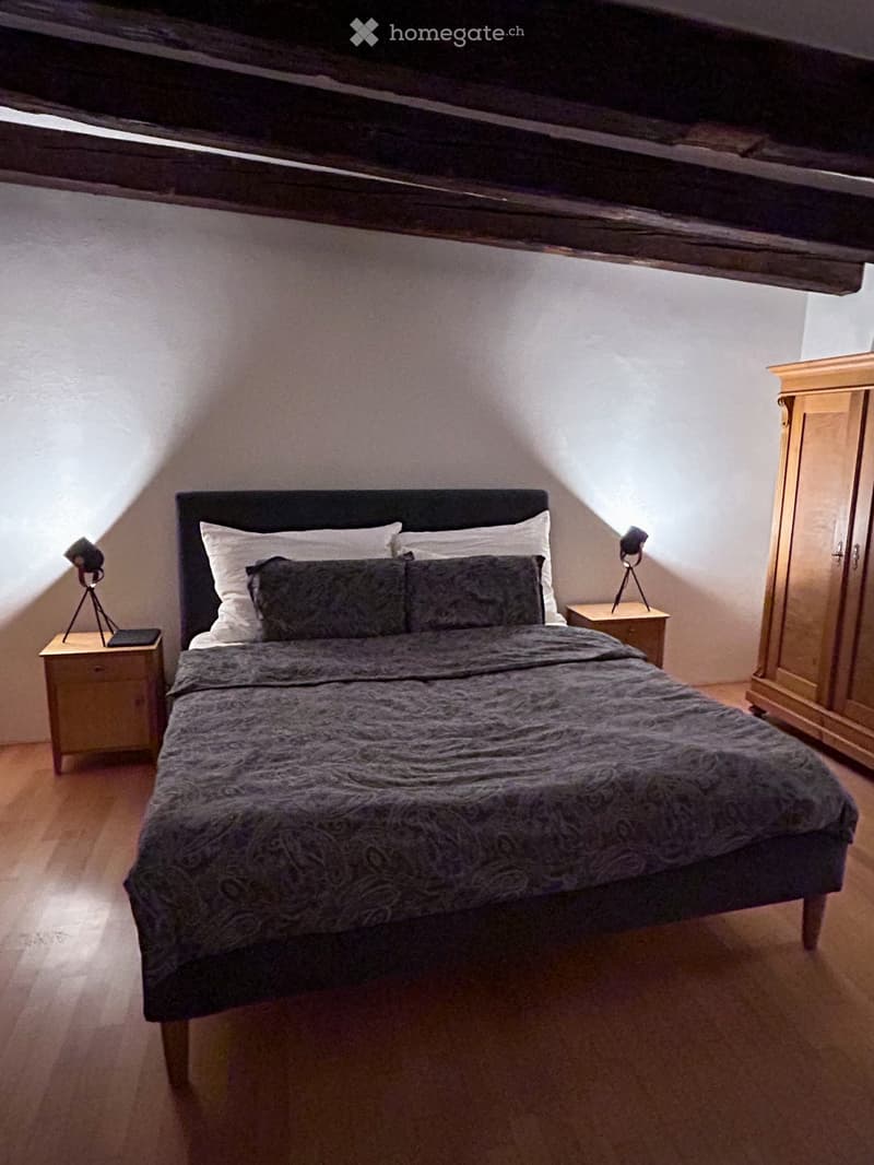 Cozy bedroom with a wooden bed, nightstands, lamps, and a wardrobe, featuring exposed wooden beams on the ceiling.