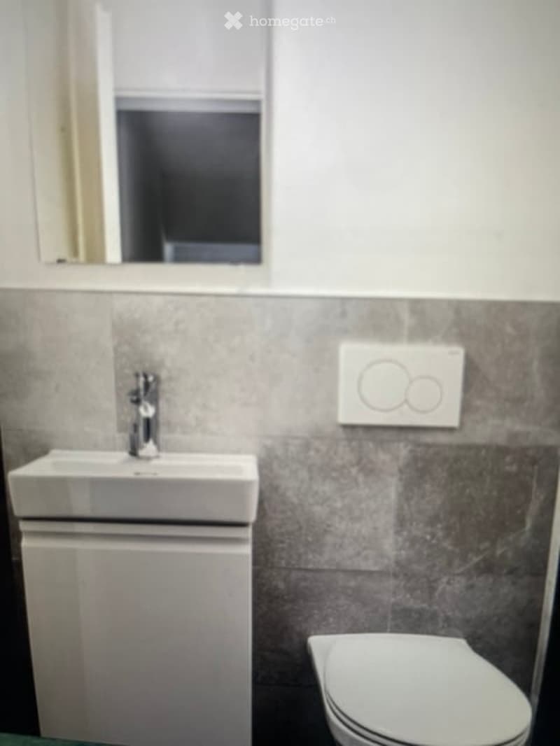 Modern bathroom with a white sink, mirror, toilet, and cabinet. Grey tiled walls and a sleek chrome faucet.