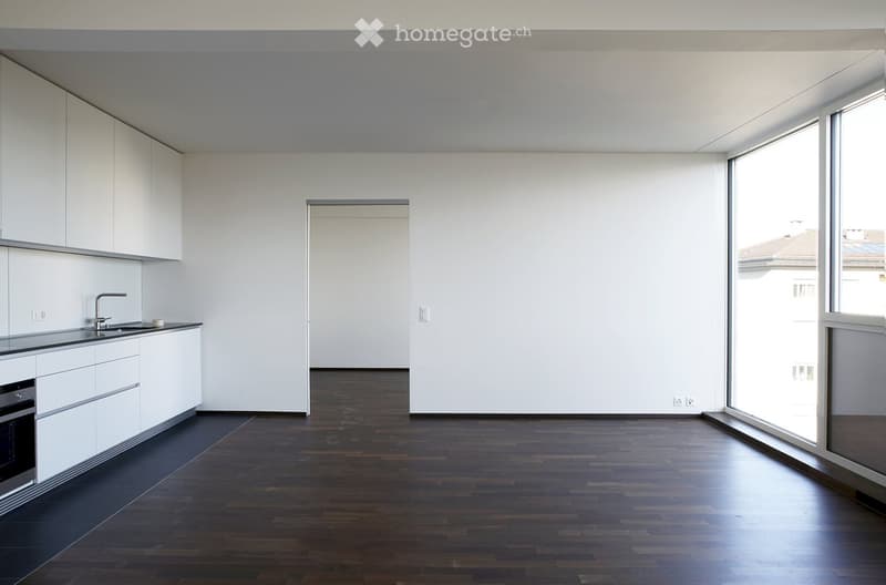 Empty kitchen with white cabinets, dark wooden floor, and large windows. No furniture or appliances are present.