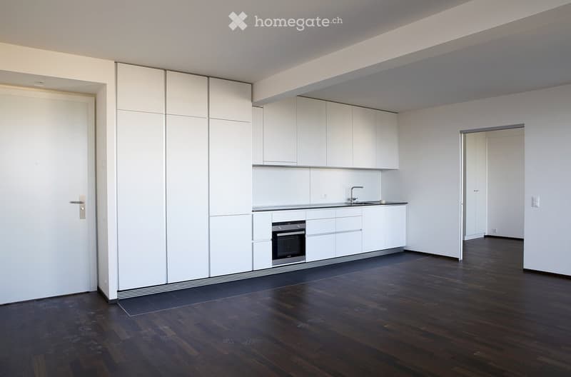Empty room with white walls, dark wooden flooring, and a small white kitchen area featuring cabinets, a sink, and an oven. The space is clean and unfurnished.