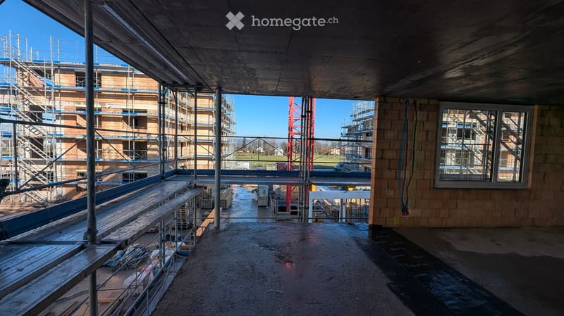 An empty, unfinished room with exposed concrete floors, wooden beams, and scaffolding visible in the background. The room is part of a larger construction site with visible construction materials and structures.