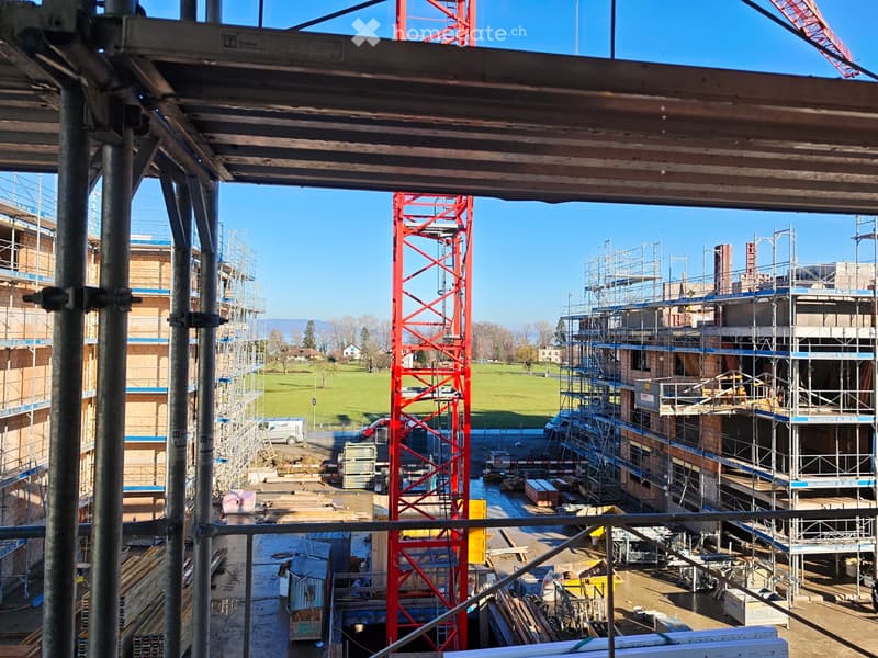 Construction site with scaffolding, cranes, and partially built structures, viewed from a high vantage point with a clear blue sky and distant park visible.