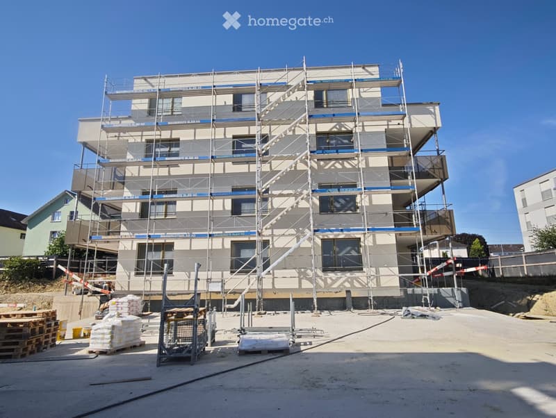 Multi-story building under construction with scaffolding, visible windows, and construction materials on the ground.