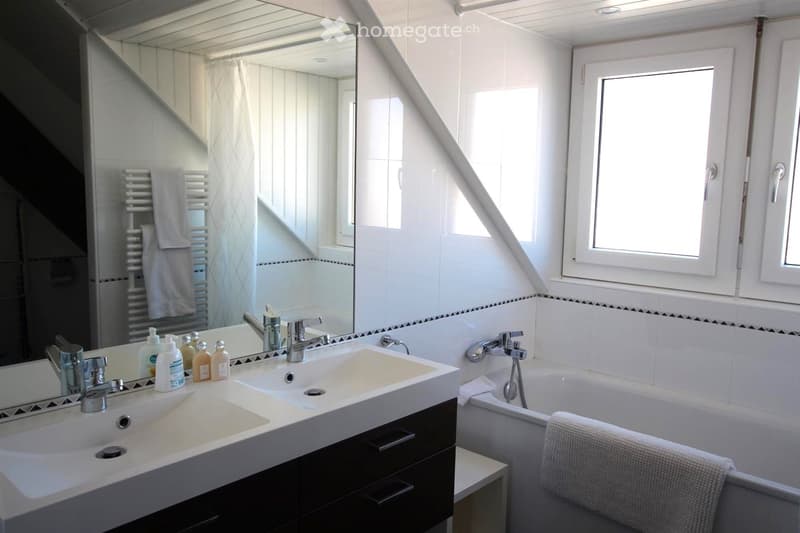 Modern bathroom with a double sink, large mirror, bathtub, and towel on the edge. The room features white tiles and a window letting in natural light.
