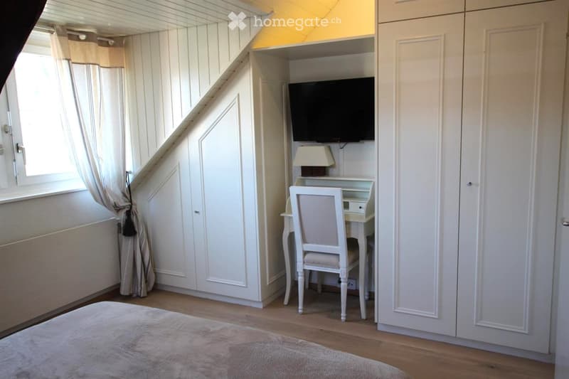 Cozy bedroom with a large window, white walls, wooden floor, and a built-in desk area with a chair, lamp, and television.