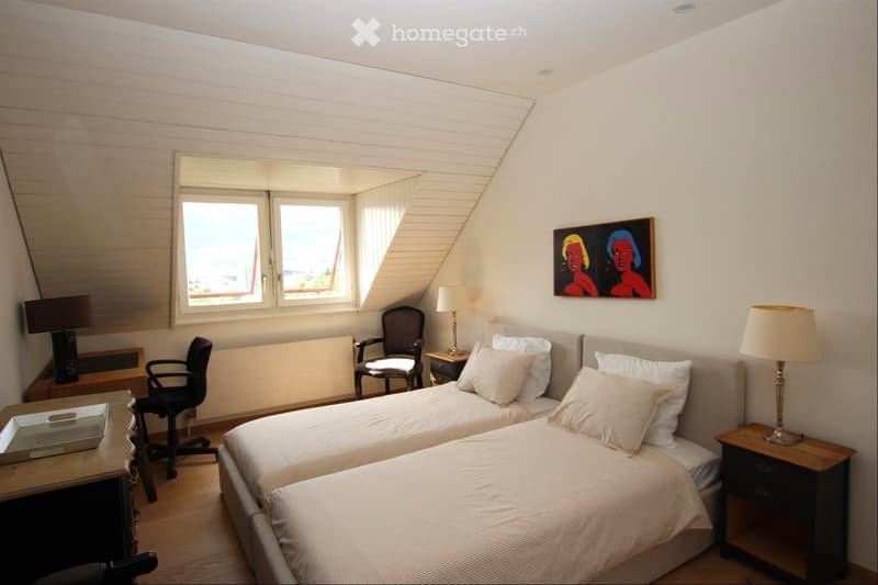 Cozy bedroom with two single beds, nightstands, lamps, a desk, and a chair, featuring large windows with a view.