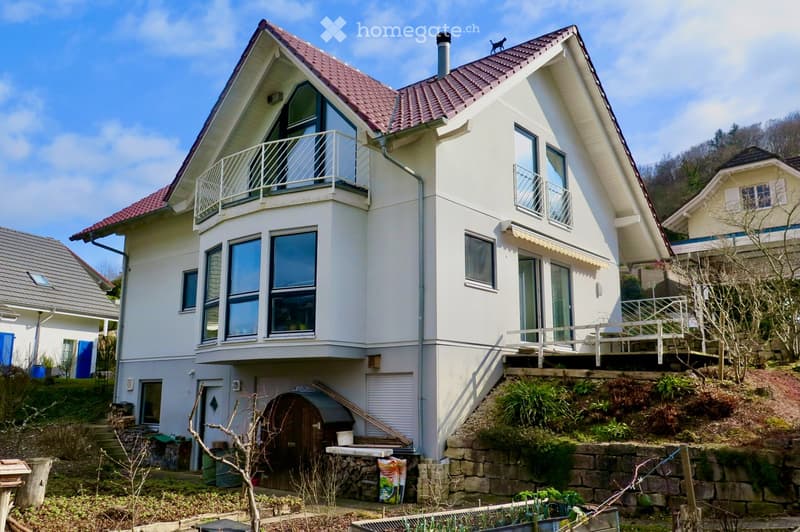 Two-story house with white exterior, red-tiled roof, multiple large windows, and a balcony on the upper level. The house is situated on a raised ground floor with a stone retaining wall and landscaped garden area.
