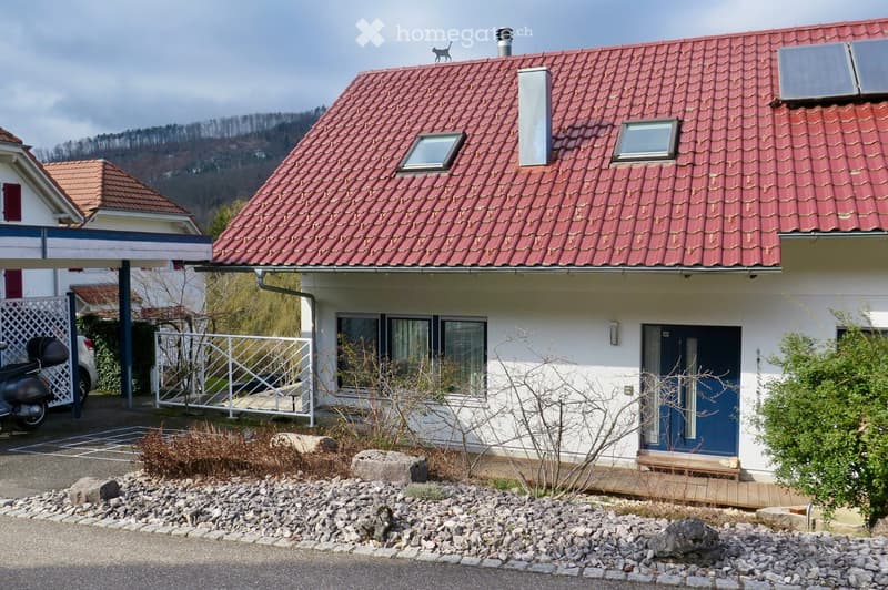 Two-story house with red-tiled roof, white walls, and blue front door. Features include a small wooden bench near the entrance, a scooter parked in the driveway, and a fenced area with a small gate.