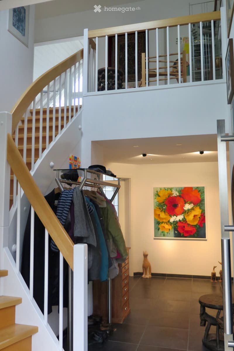 Spacious hallway with a wooden staircase leading to an upper level, coat rack with jackets, artwork on the wall, and a small table with a decorative item.