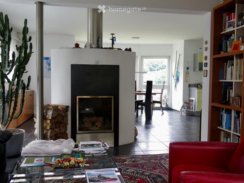 Modern living room with a red leather sofa, glass coffee table, bookshelf, dining area with chairs and table, and a fireplace with wood stack nearby.