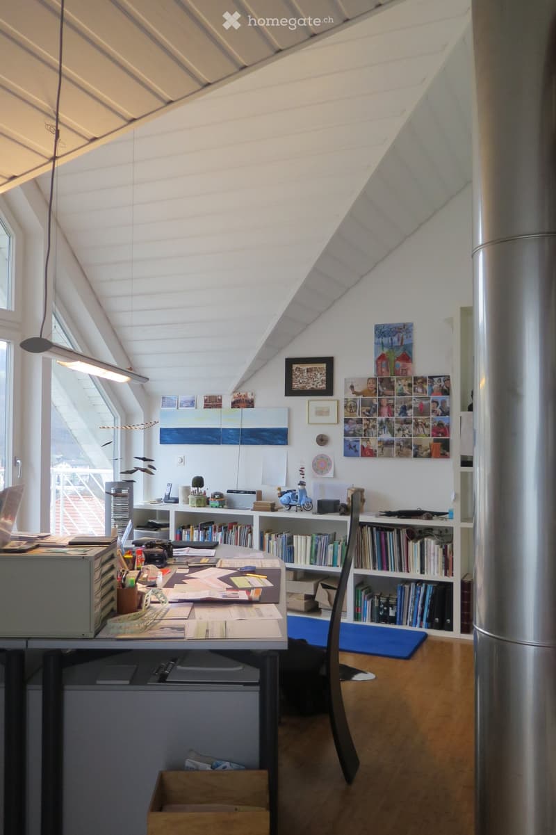Bright, airy attic room with slanted ceilings, large windows, and modern furnishings including a desk, bookshelf, and artwork.