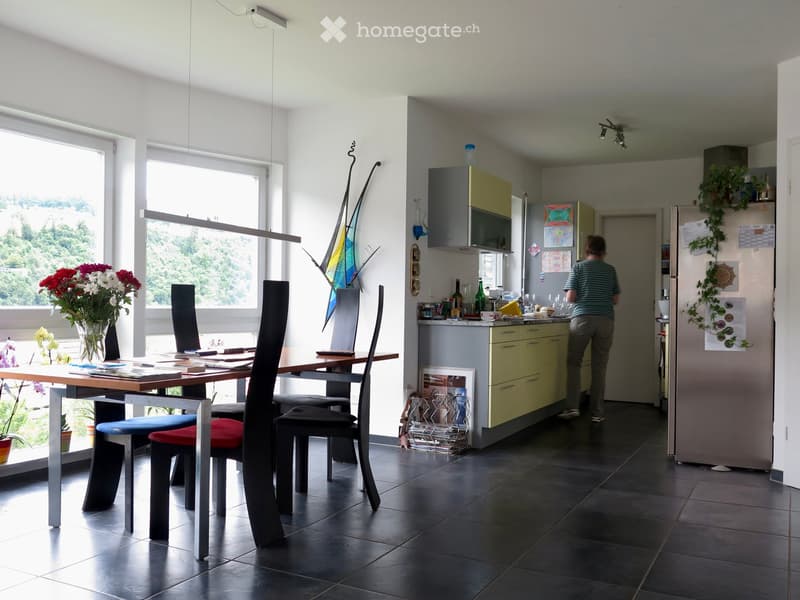 A modern, open-plan kitchen and dining area with a large window offering a scenic view. The dining area features a wooden table with four contemporary chairs, a vase with flowers, and decorative items. The kitchen includes modern cabinets, a refrigerator,