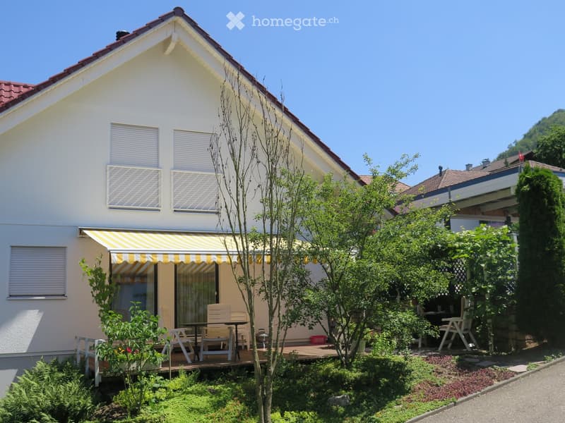 Two-story house with white exterior, red-tiled roof, and striped awning over the entrance. The front yard features a wooden deck with outdoor furniture, including chairs and a table, surrounded by greenery and plants.