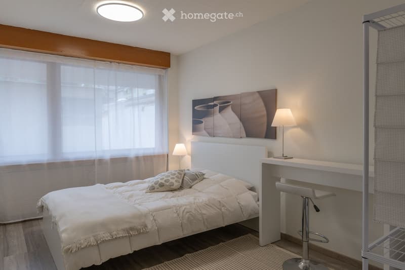 A modern, well-lit bedroom with a large bed, decorative pillows, a white headboard, a small desk with a stool chair, and a wall-mounted picture of vases.
