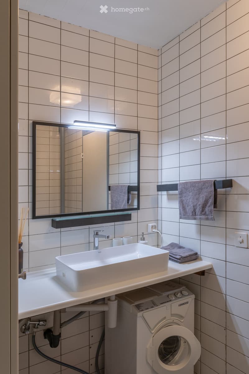 Modern bathroom with white tiles, a large sink, a mirror with built-in lighting, and a washing machine underneath the sink.