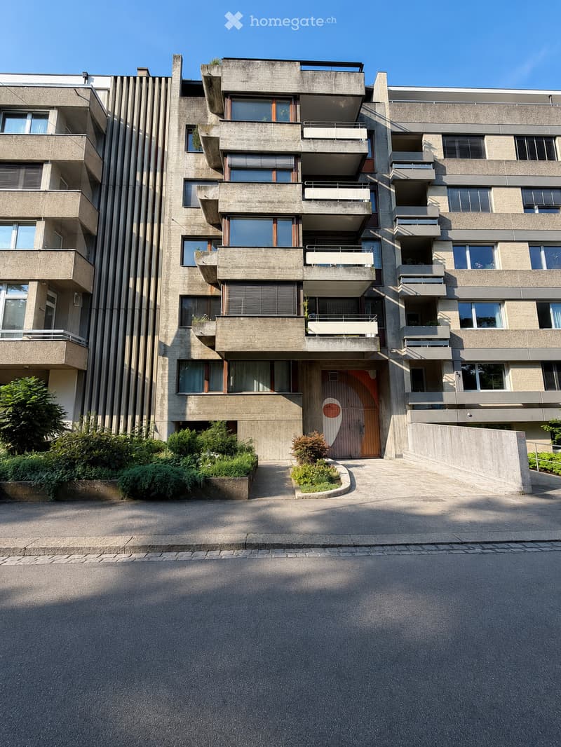 Modern multi-story apartment building with concrete facade, large windows, and balconies. Features a central entrance with a distinctive circular design above the door.