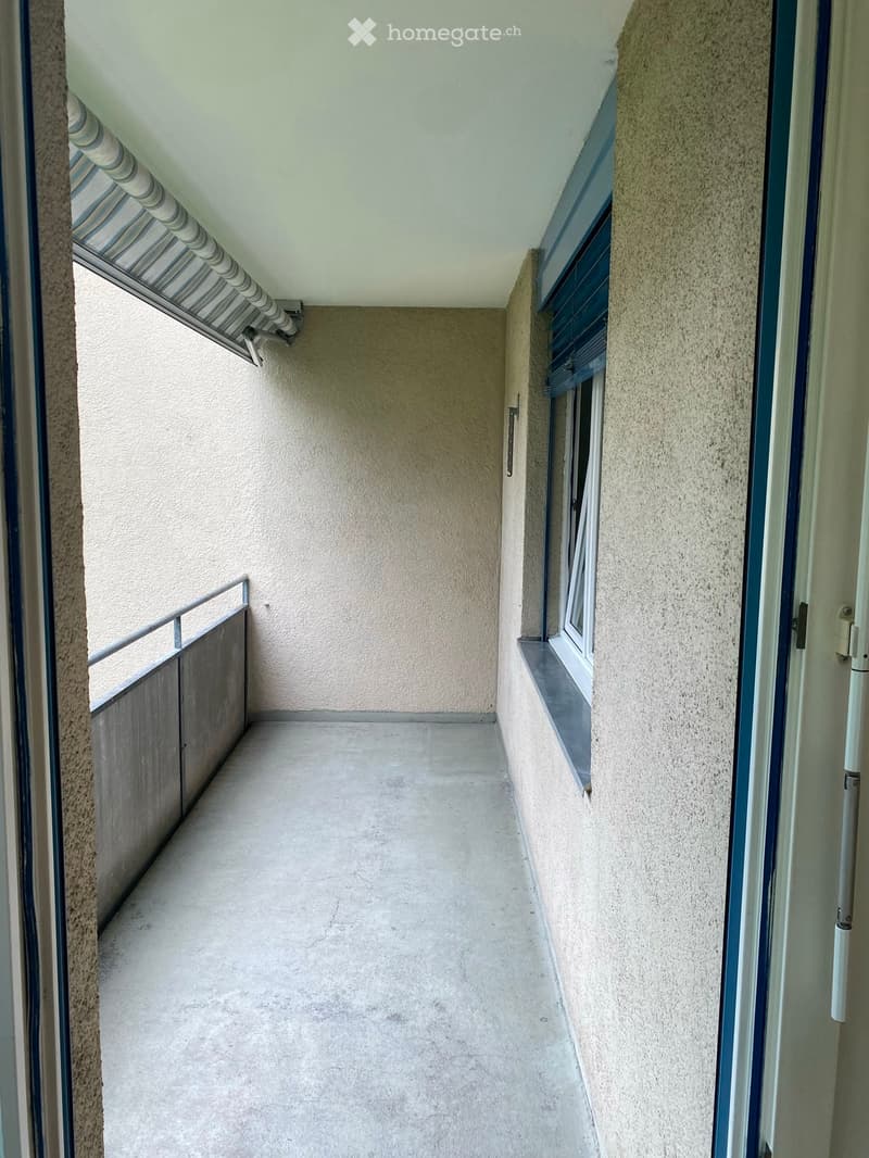 Empty balcony with concrete floor, beige walls, metal railing, and a window with shutters.