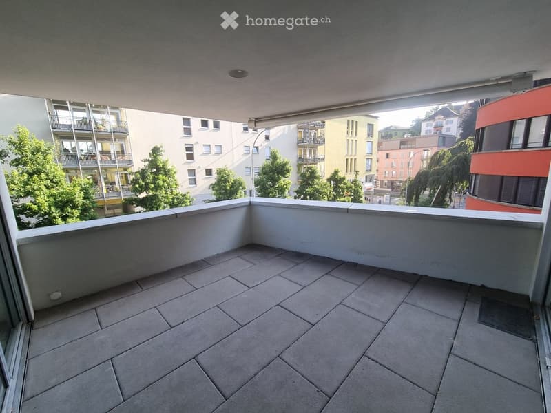 Empty balcony with tiled floor, white railing, and a view of surrounding buildings and greenery.