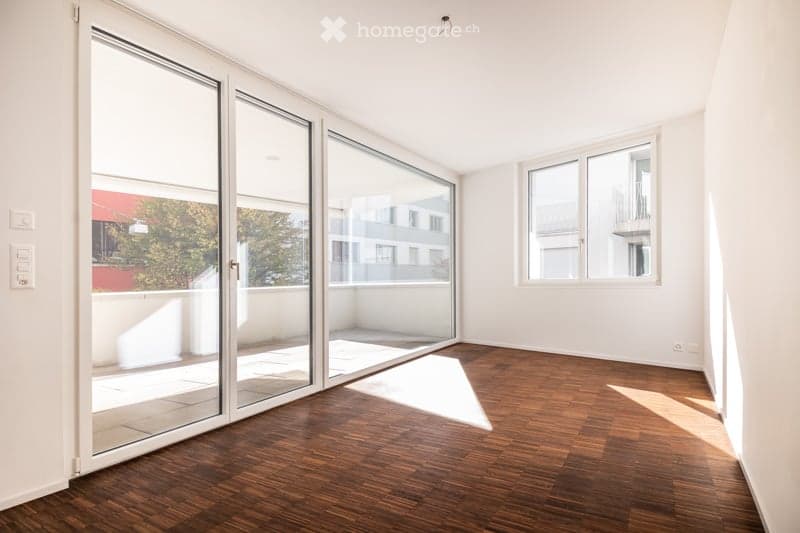 Bright, unfurnished room with wooden flooring, large sliding glass doors leading to a balcony, and ample natural light.
