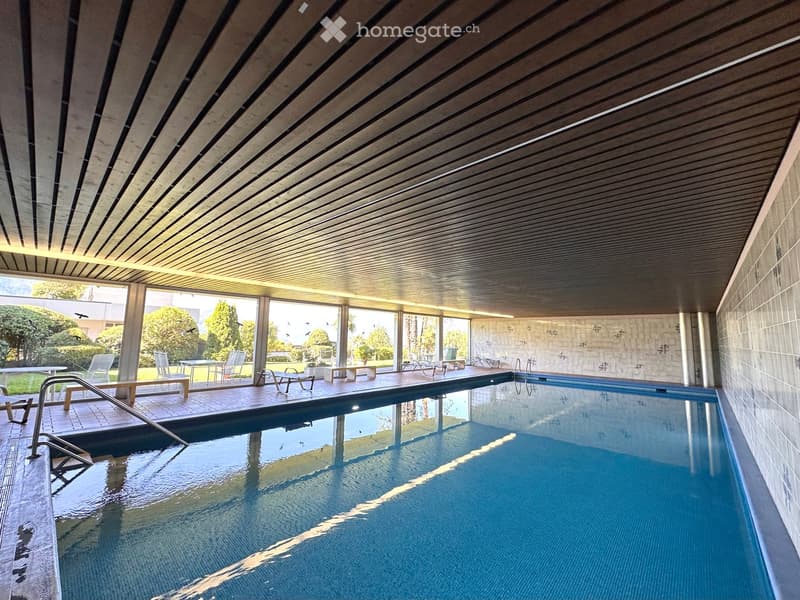 Indoor pool area with large windows, surrounded by lounge chairs and tables, with a view of the outdoor garden and trees.