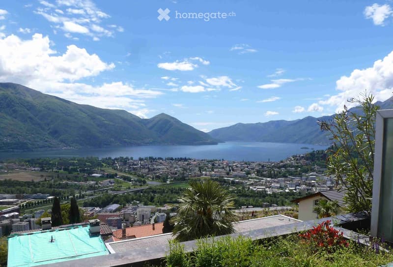 Panoramic view from a balcony or terrace overlooking a town, lake, and surrounding mountains under a partly cloudy sky.