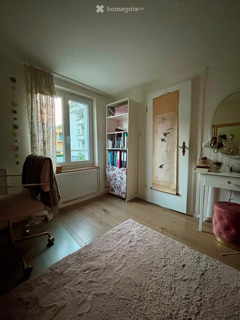 A furnished room with a wooden floor, a large window with curtains, a bookshelf, a chair, a mirror, a small table, and a pink stool.