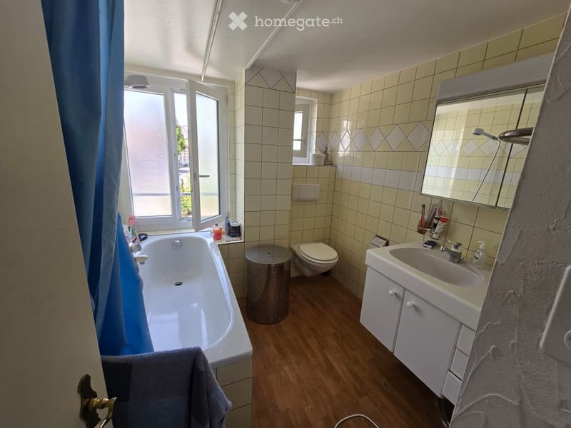Bathroom with a bathtub, toilet, sink, mirror, and cabinet. The room has tiled walls, wooden flooring, and a window with a blue curtain.