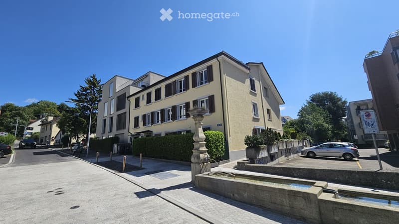 Multi-story residential building with beige facade, multiple windows with shutters, and a small fountain in front.