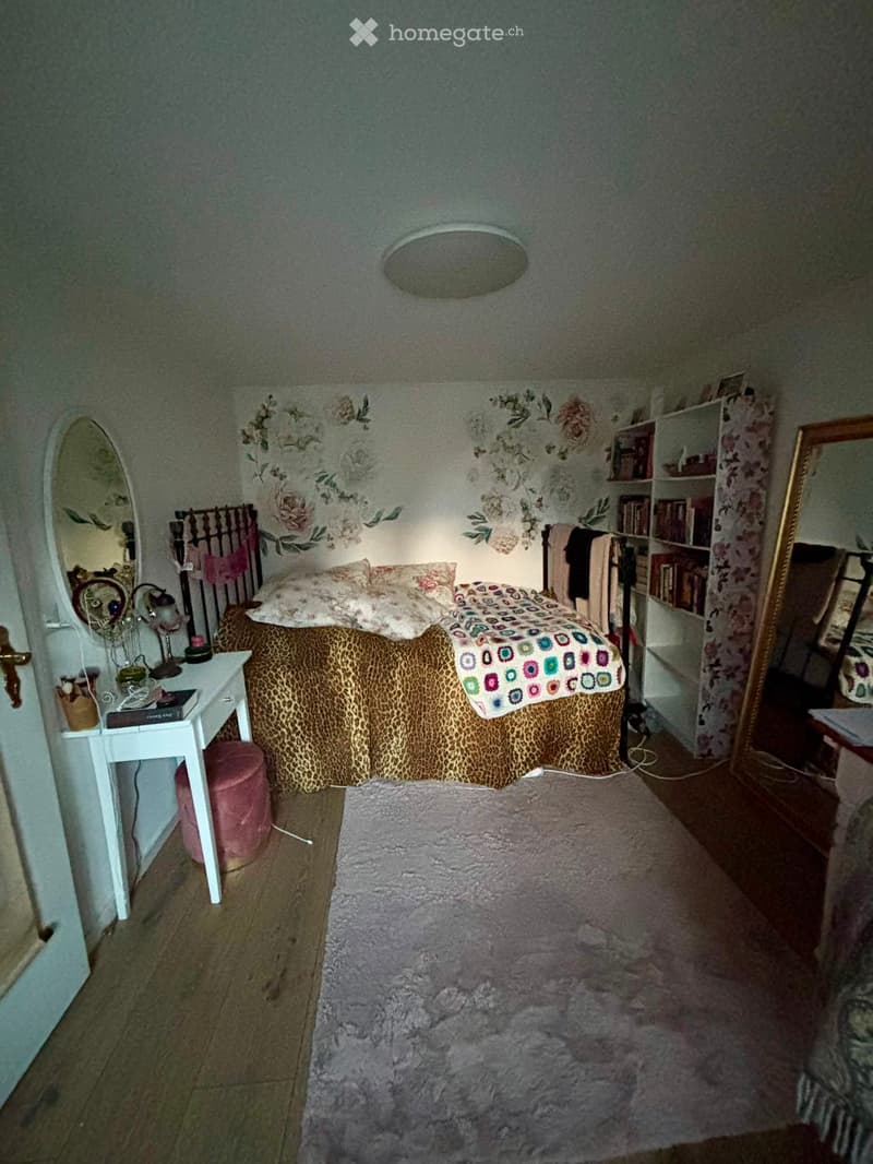 Cozy bedroom with a wooden floor, floral wallpaper, a large bed with leopard print bedding, a fluffy rug, a white nightstand, a pink stool, and a bookshelf.