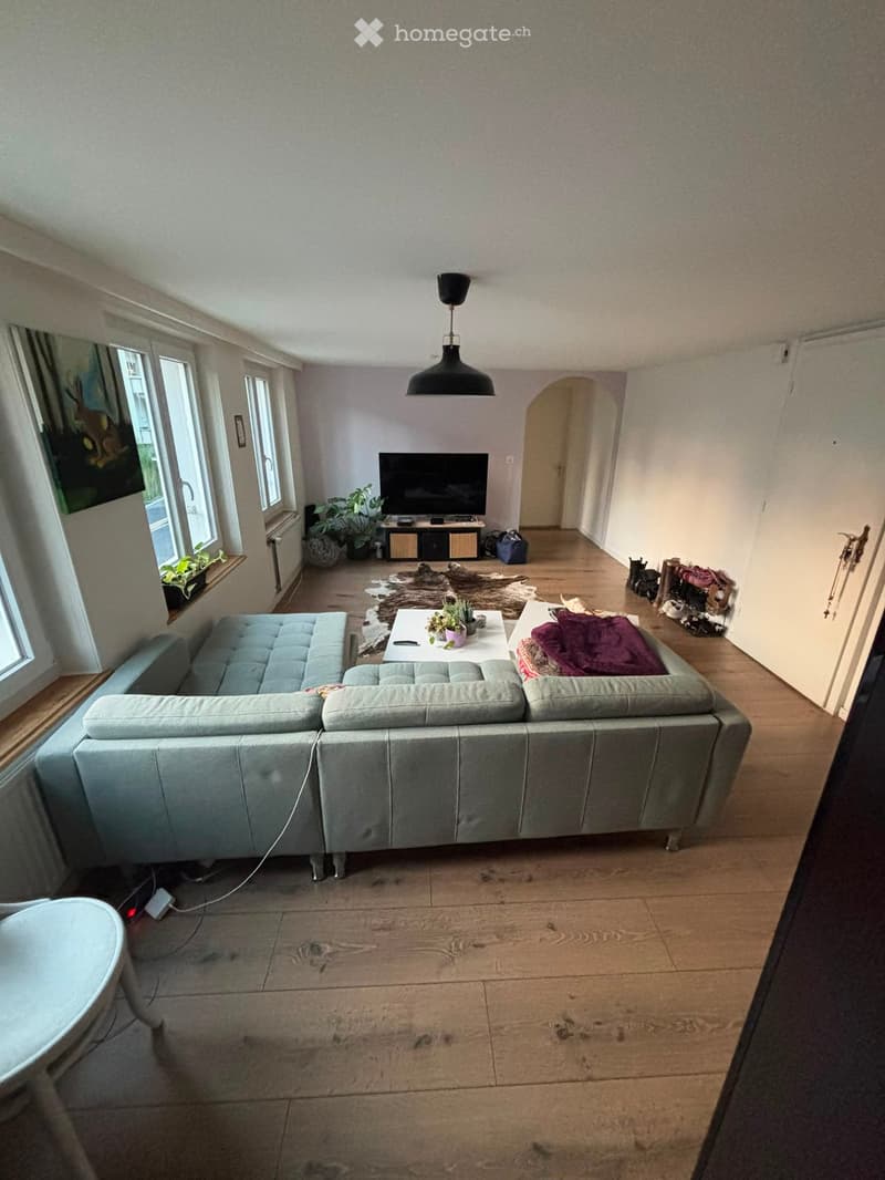 Modern living room with light grey sectional sofa, coffee table, TV, floor lamp, and plants. Wooden flooring and large windows.