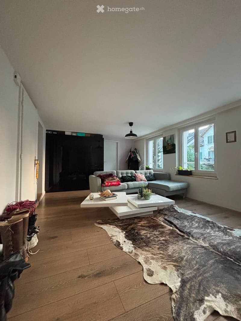 Living room with wooden flooring, a grey sectional sofa, a white coffee table, a cowhide rug, and a potted plant on the windowsill.