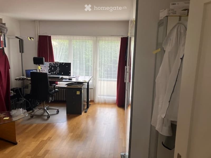 A furnished room with a wooden floor, large window with curtains, a desk with a computer and screen, a chair, and a lamp.