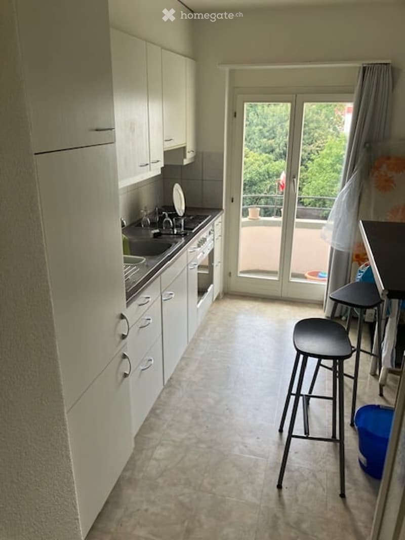 Modern kitchen with white cabinets, stainless steel sink, oven, and two black stools near a small table. A sliding glass door leads to a balcony with outdoor furniture.