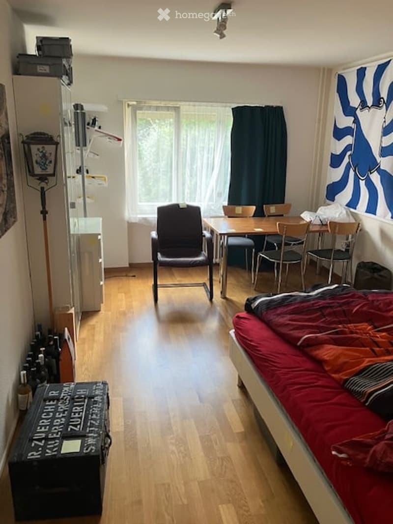 A small, furnished room with a bed, a table, chairs, a bookshelf, and a curtain. The room has wooden flooring and large windows letting in natural light.