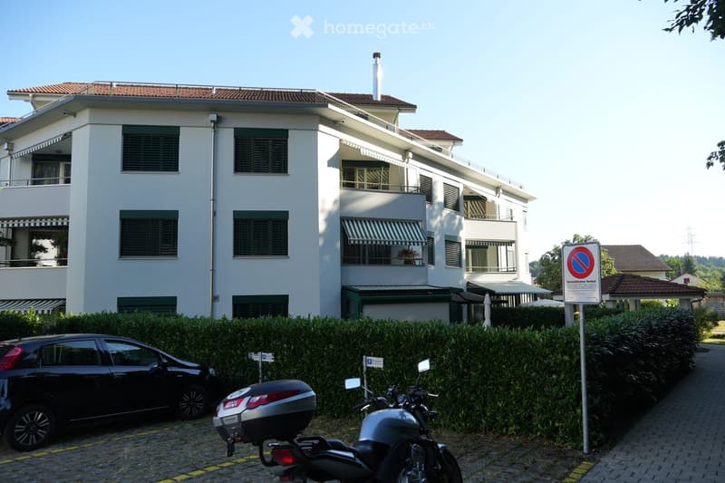 A modern, multi-story residential building with white walls, green shutters, and balconies. The building features a red-tiled roof and is surrounded by a neatly trimmed hedge. A black car and a motorcycle are parked in front of the building.