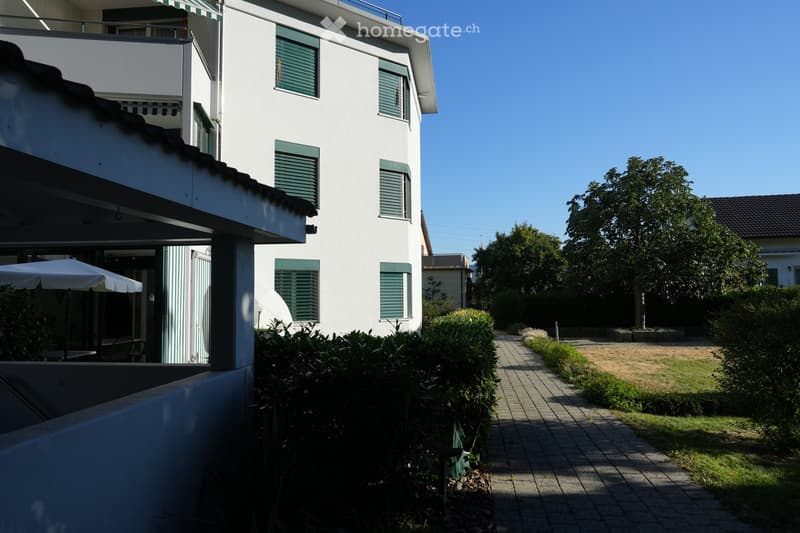 A modern white building with green shutters and balconies, surrounded by a well-maintained garden with a paved pathway, greenery, and an umbrella on the balcony.