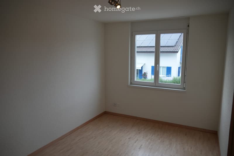 Empty room with wooden flooring, white walls, and a large window with blinds. The room is unfurnished and has a simple, clean design.