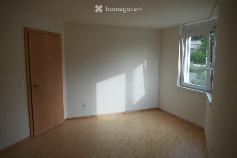 Empty room with wooden flooring, white walls, and a single window letting in natural light. The room features a wooden door on one side and electrical outlets on the walls.