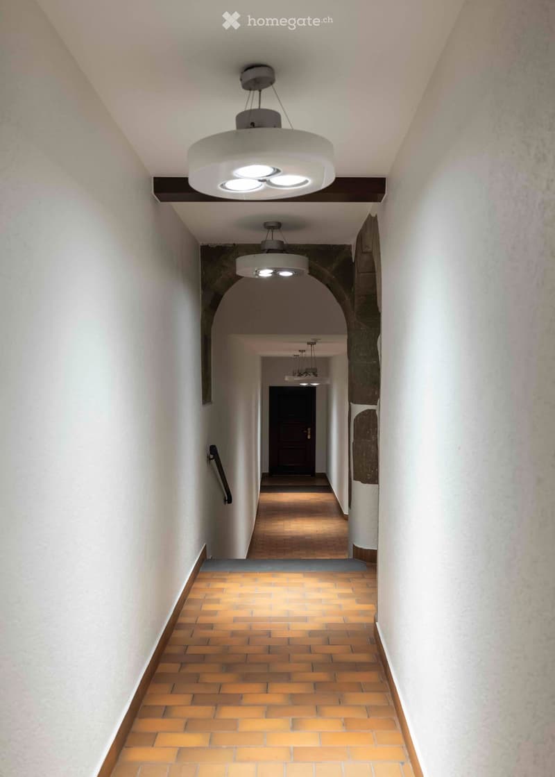 Staircase with wooden steps and brick tiles, modern pendant lights, white walls, and a dark wooden door at the end.