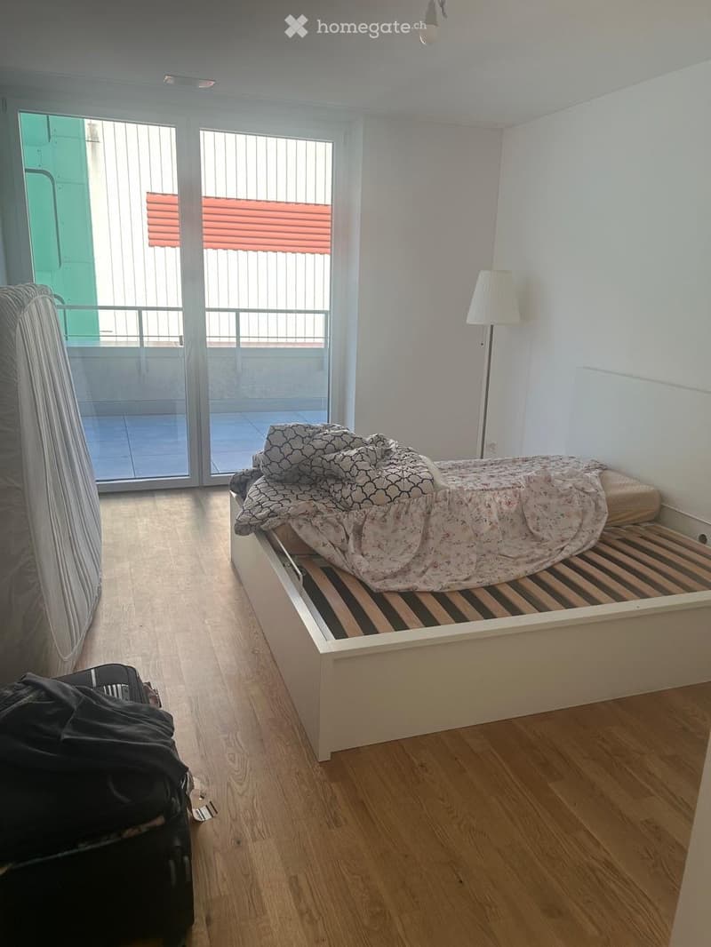 Bedroom with wooden floor, bed with patterned bedding, floor lamp, suitcase, and large window with blinds.