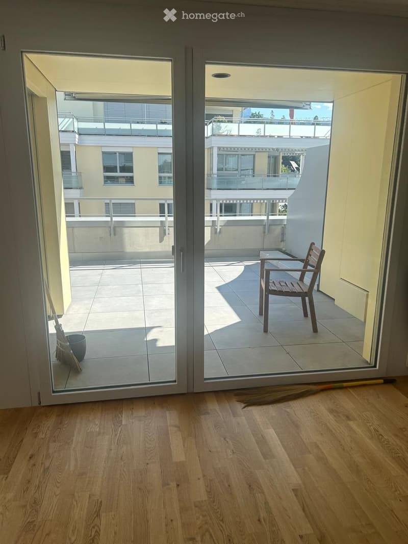 Empty room with wooden flooring and large sliding glass doors leading to a tiled balcony area. The balcony has a wooden chair and a broom is visible near the doors.