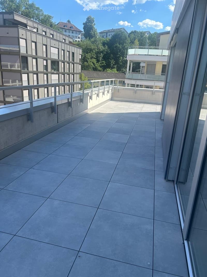 Clean, modern balcony with tiled floor and metal railing, overlooking a cityscape with buildings and greenery.