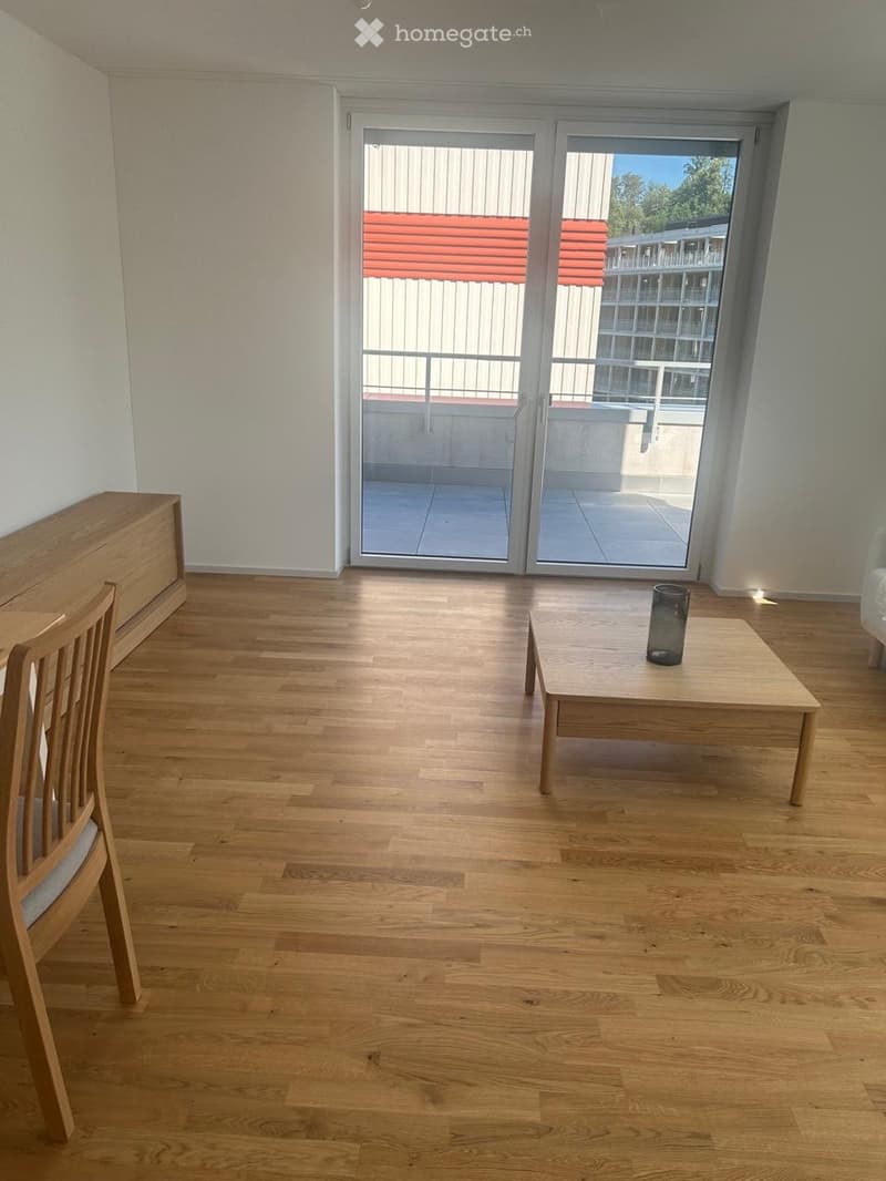 A modern, unfurnished living room with wooden flooring, a sliding glass door leading to a balcony, a wooden chair, a small table with a cup, and a wooden cabinet.