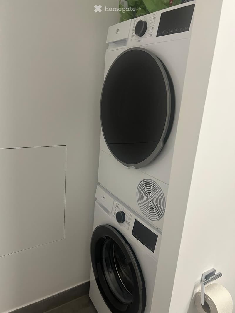 A compact bathroom with a stacked washing machine and dryer unit, white cabinetry, and a toilet paper holder.
